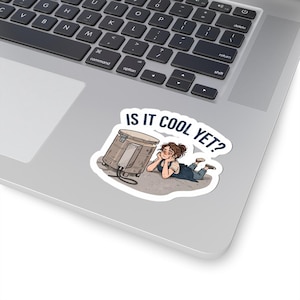 May include: A die-cut sticker featuring a cartoon illustration of a person lying on the ground, resting their chin on their hands while looking at a cylindrical kiln. The text above the illustration reads IS IT COOL YET? in dark blue letters.