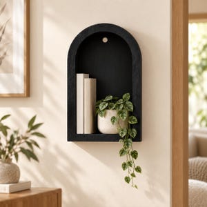 Puede incluir: Un estante de pared arqueado de color negro con acabado liso y un orificio circular de montaje en la parte superior. Dentro del estante hay dos libros beige verticales y una maceta de cerámica blanquecina con una planta verde colgante.