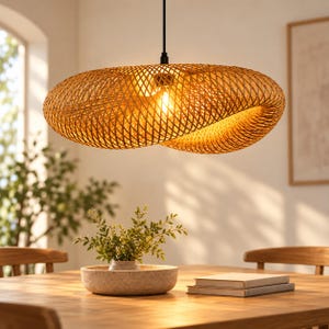 Può includere: Una grande lampada a sospensione di forma ovale realizzata con strisce di bambù intrecciate color marrone chiaro. Il paralume presenta un intricato motivo a reticolo incrociato che diffonde una luce calda attraverso le fessure, sospeso da un cavo nero.
