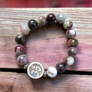 May include: A bracelet made of pink and grey gemstone beads with a white ceramic charm featuring an Om symbol.
