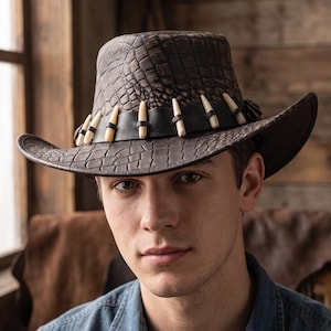May include: A dark brown hat with a textured crocodile skin pattern. The hat features a wide brim and a black leather band adorned with a series of off-white, pointed tooth-like ornaments spaced evenly around the crown of the headwear.