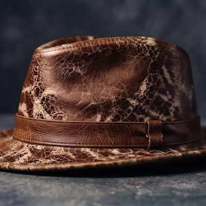 May include: A brown fedora hat featuring a distinct cracked, distressed texture across the crown and brim. The hat includes a smooth, solid brown leather band encircling the base of the crown, providing a contrast to the weathered finish.