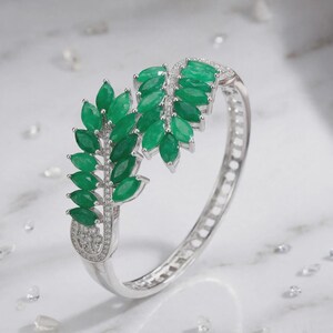 May include: A silver-toned hinged bangle bracelet featuring two leaf-shaped motifs adorned with marquise-cut green stones and small clear accent stones. The metal band has a cut-out design along the side and a polished finish.