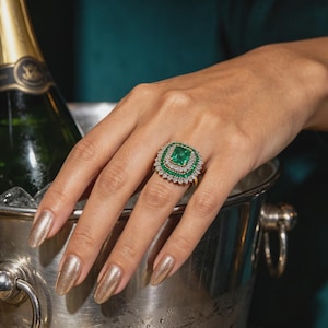 May include: A gold ring featuring a large rectangular emerald centre stone surrounded by a double halo of smaller round emeralds and diamonds. The ring has a wide, ornate setting with a polished gold band, showcasing intricate metalwork details.