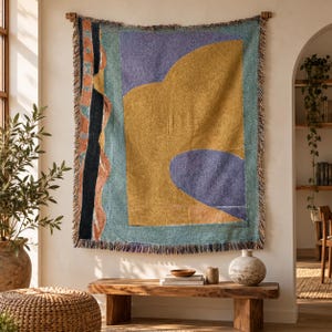 Pode incluir: Uma tapeçaria de parede tecida retangular com um design abstrato apresentando grandes blocos de amarelo mostarda, roxo suave e verde-azulado. O lado esquerdo exibe uma faixa preta vertical com bordas estampadas em laranja e azul.
