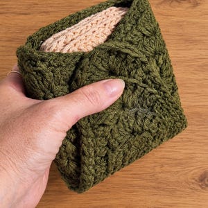 May include: A square, olive green crocheted pouch featuring a textured, repeating stitch pattern. The pouch is folded over at the top, revealing a soft, light peach-colored fabric lining tucked inside the dark green exterior.