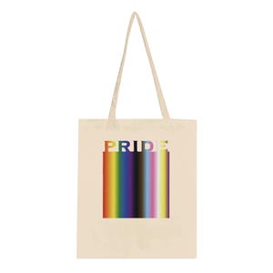 Op de afbeelding: Een beige canvas draagtas met twee lange schouderbanden. Het midden van de tas is voorzien van een rechthoekige afbeelding met een verticaal regenboogverloop en het woord PRIDE in witte hoofdletters boven op het gekleurde blok.