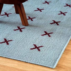 May include: A light blue area rug featuring a repeating pattern of dark red cross symbols. The rug has a textured, woven appearance and is partially covered by the wooden legs of a piece of furniture, showing a consistent grid-like design.