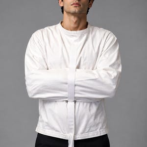 May include: A long-sleeved white shirt featuring a high collar and a vertical white strap running down the center front, secured with a metal ring at the waist. The garment has long sleeves that are folded across the chest and black trousers.