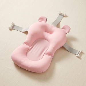 Puede incluir: Un cojín de baño suave de color rosa claro con una forma contorneada y dos protuberancias redondeadas similares a orejas en la parte superior. Incluye tres correas grises ajustables con clips de plástico blanco fijados a los lados y arriba.