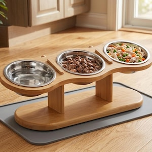 Puede incluir: Una estación de alimentación elevada para mascotas con tres cuencos de acero inoxidable extraíbles en un soporte de madera de bambú clara. Los cuencos contienen agua, trozos de carne marrón y una mezcla de pollo con guisantes y zanahorias.