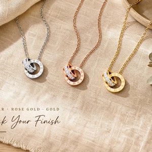 Puede incluir: Tres collares en tonos plata, oro rosa y oro, cada uno con un colgante de dos anillos entrelazados. Un anillo está grabado con números romanos, mientras que el otro tiene pequeñas piedras claras. Texto: SILVER ROSE GOLD GOLD Pick Your Finish.