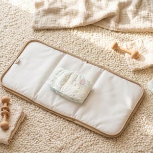 Puede incluir: Un cambiador rectangular blanco con borde beige, dos pañales plegados, dos sonajeros de madera, una pequeña cesta tejida y un paño de muselina beige doblado. Los artículos están dispuestos sobre una superficie texturizada de color crema.