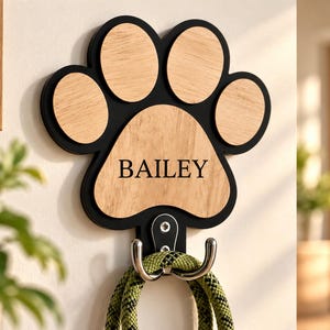Puede incluir: Un gancho de pared con forma de huella con un marco negro e inserciones de madera clara. La almohadilla central está grabada con el nombre BAILEY en letras mayúsculas negras. Un gancho doble de metal plateado sostiene una correa de cuerda trenzada verde.