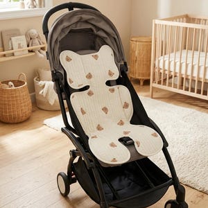 May include: A black stroller featuring a cream-colored seat liner with a repeating brown bear pattern. The liner has a contoured shape with ear-like top sections, a central cutout for harness straps, and a quilted texture throughout the fabric.