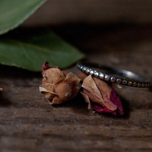 Può includere: Un anello d'argento con un designo a perline si trova su una superficie di legno con boccioli di rosa secchi.