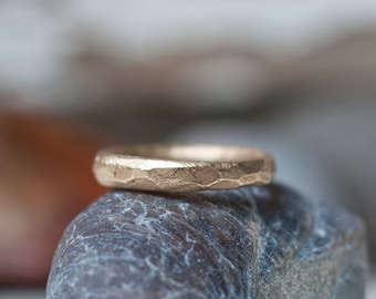 May include: A gold hammered ring sits on top of a gray rock. The ring is a simple band with a textured surface.