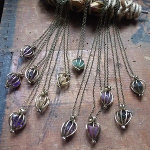 Könnte beinhalten: Eine Sammlung von 12 Halsketten mit Käfig-Kristallanhängern. Jede Halskette hat einen andersfarbigen Kristall, darunter Amethyst, Grün und Klar.