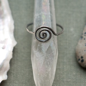 Könnte beinhalten: Ein silberfarbener Ring mit einem Spiraldesign. Der Ring wird auf einem klaren, kristallartigen Objekt präsentiert, mit einer Muschel und einem gesprenkelten Stein im Hintergrund. Die Spirale steht im Mittelpunkt und erzeugt eine minimalistische und elegante Ästhetik.