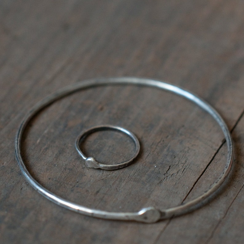 Op de afbeelding: Twee zilveren ringen, een groter dan de andere, liggen op een houten oppervlak. De grotere ring is een eenvoudige cirkel, terwijl de kleinere ring een lichte kromming in zijn vorm heeft.