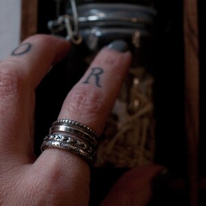 Puede incluir: Una mano con una pila de cuatro anillos de plata. Los anillos son de diferentes estilos, algunos con un acabado texturizado. La mano est&aacute; llegando a un frasco de vidrio.