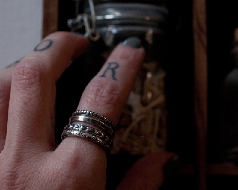 Puede incluir: Una mano con una pila de cuatro anillos de plata. Los anillos son de diferentes estilos, algunos con un acabado texturizado. La mano est&aacute; llegando a un frasco de vidrio.