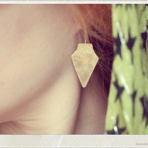 May include: A close-up of a person's ear with a gold-coloured arrow-shaped earring. The earring is a simple design with a pointed tip and a slightly rounded base.