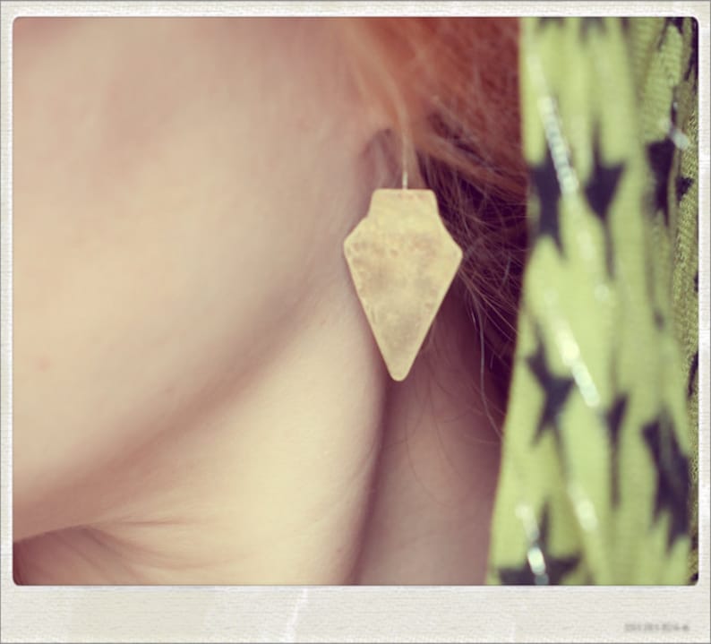 May include: A close-up of a person's ear with a gold-coloured arrow-shaped earring. The earring is a simple design with a pointed tip and a slightly rounded base.