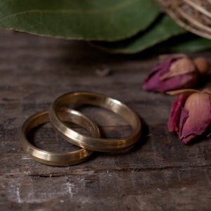 Puede incluir: Dos anillos de boda dorados, entrelazados, descansan sobre una superficie de madera rústica. Los anillos tienen un diseño simple y clásico. Capullos de rosa secos y hojas verdes en el fondo, añadiendo un toque de belleza natural a la escena.