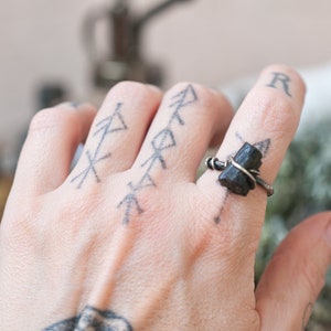 May include: A silver ring with a black tourmaline gemstone wrapped in wire. The ring is on a person's finger.