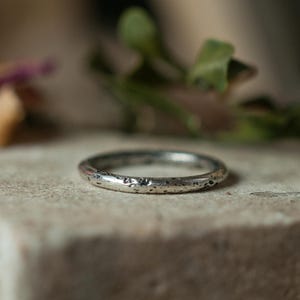 Könnte beinhalten: Ein silberner Ring mit strukturierter Oberfläche. Der Ring ist kreisförmig und scheint aus Metall zu sein. Der Ring liegt auf einer neutralfarbenen Oberfläche mit verschwommenem Grün im Hintergrund.
