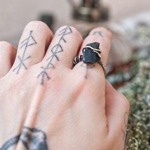 May include: A silver ring with a black stone wrapped in wire. The ring is on a person's finger with a tattoo of several arrows.