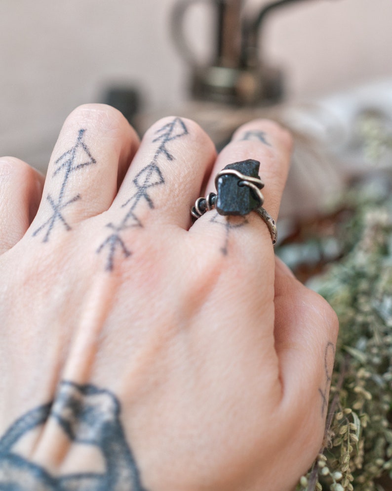May include: A silver ring with a black stone wrapped in wire. The ring is on a person's finger with a tattoo of several arrows.
