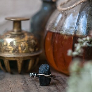 May include: A black stone ring with a silver wire wrap. The ring is sitting on a wooden surface with a glass jar and a brass bell in the background.