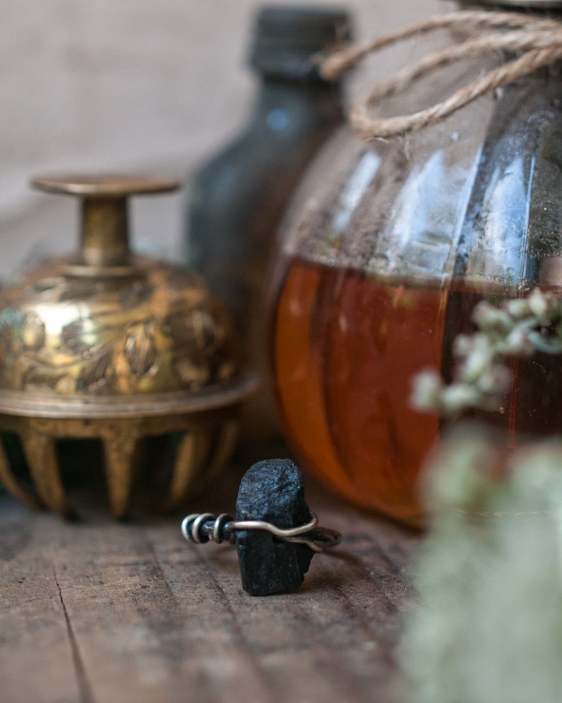 May include: A black stone ring with a silver wire wrap. The ring is sitting on a wooden surface with a glass jar and a brass bell in the background.