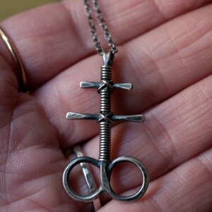 K&ouml;nnte beinhalten: Eine silberne Halskette mit einem gewundenen, gedrehten Anh&auml;nger, der ein Kreuz und einen Kreis zeigt. Der Anh&auml;nger ist aus Metall und hat eine dunkle, gealterte Oberfl&auml;che.