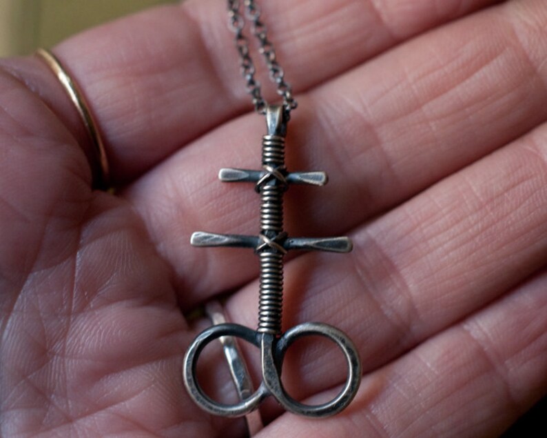 K&ouml;nnte beinhalten: Eine silberne Halskette mit einem gewundenen, gedrehten Anh&auml;nger, der ein Kreuz und einen Kreis zeigt. Der Anh&auml;nger ist aus Metall und hat eine dunkle, gealterte Oberfl&auml;che.