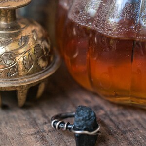 May include: A black gemstone ring with a silver band. The ring is set on a wooden surface with a gold bell and a glass jar in the background.