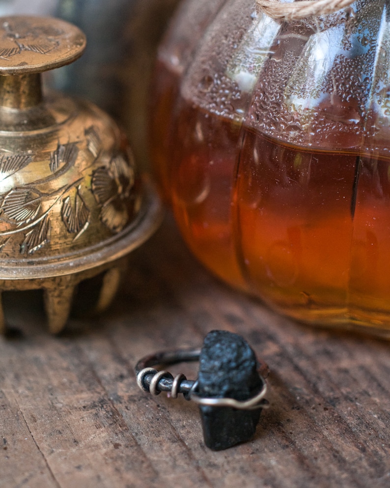 May include: A black gemstone ring with a silver band. The ring is set on a wooden surface with a gold bell and a glass jar in the background.