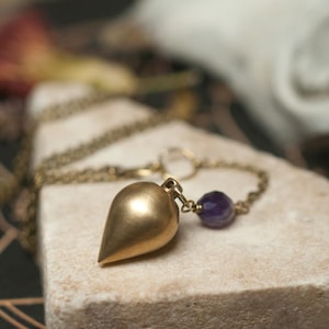 May include: A teardrop-shaped brass pendant with a matte finish attached to a metallic chain. A small, faceted, deep purple amethyst bead is positioned on the chain directly next to the pendant, which features a smooth, pointed bottom.