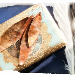Puede incluir: Un par de pendientes de cobre con un diseño en capas con forma de pluma. Los pendientes se muestran en una caja azul y dorada con un patrón floral.