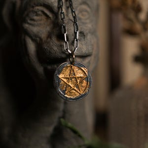 Può includere: Un ciondolo dorato con un design a stella a cinque punte, incastonato in una cornice circolare color argento. Il ciondolo è appeso a una catena d'argento. Lo sfondo presenta una scultura in pietra sfocata e usurata.