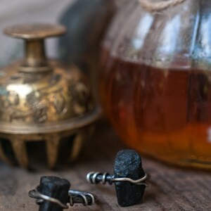 May include: Two black tourmaline rings with silver wire wrapped around the stones. The rings are on a wooden surface with a glass jar and a brass bell in the background.