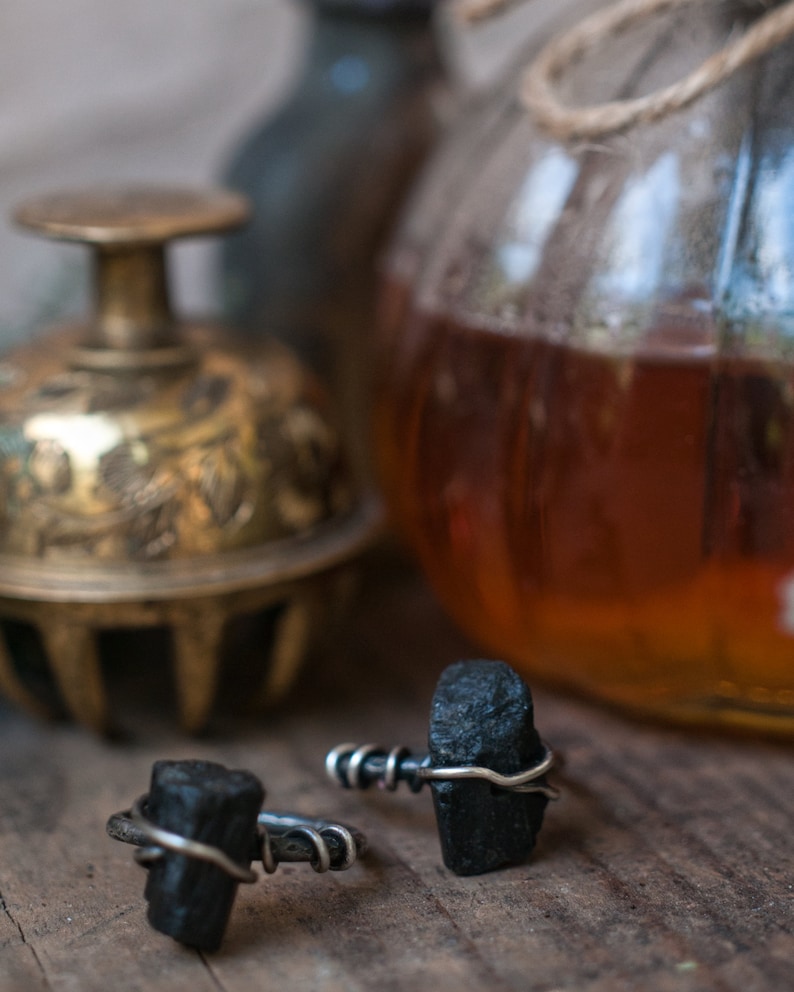 May include: Two black tourmaline rings with silver wire wrapped around the stones. The rings are on a wooden surface with a glass jar and a brass bell in the background.