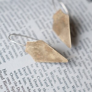 May include: A pair of silver earrings with a geometric, triangular shape. The earrings have a hammered, textured finish and a rustic, antique look.