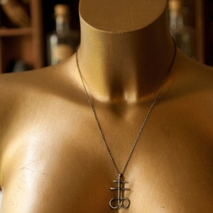 K&ouml;nnte beinhalten: Eine silberne Halskette mit einem Anh&auml;nger, der ein stilisiertes Kreuz mit zwei Kreisen zeigt. Die Halskette befindet sich auf einer goldenen Schaufensterpuppe.