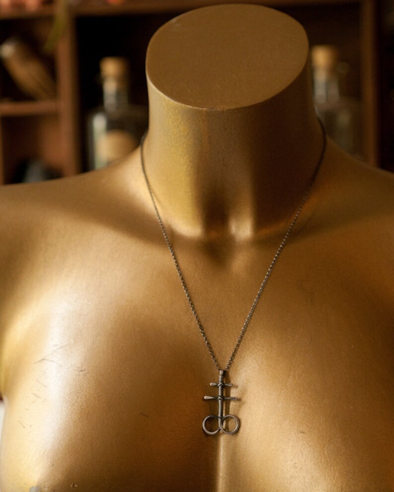 K&ouml;nnte beinhalten: Eine silberne Halskette mit einem Anh&auml;nger, der ein stilisiertes Kreuz mit zwei Kreisen zeigt. Die Halskette befindet sich auf einer goldenen Schaufensterpuppe.