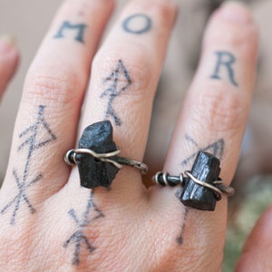 May include: Two black tourmaline rings with silver wire wrapped around the stones. The rings are worn on the index and middle fingers of a hand with tattoos.