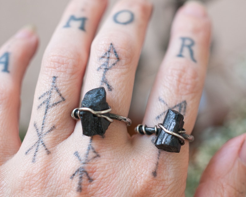 May include: Two black tourmaline rings with silver wire wrapped around the stones. The rings are worn on the index and middle fingers of a hand with tattoos.