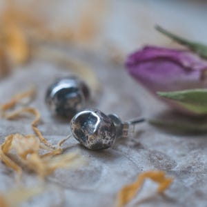 May include: A pair of silver stud earrings with a textured, irregular, pebble-like design. The earrings are on a white surface with yellow flower petals.